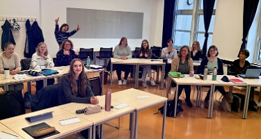 Klassenfoto: Mehrere Personen sitzen an Tischen in einem Klassenzimmer und schauen zur Kamera