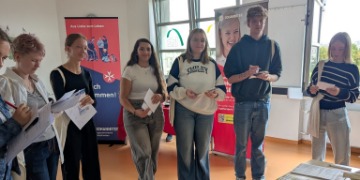 Jobbörse Schüler*innen stehen in einem Klassenzimmer und halten Informationsmaterial in den Händen